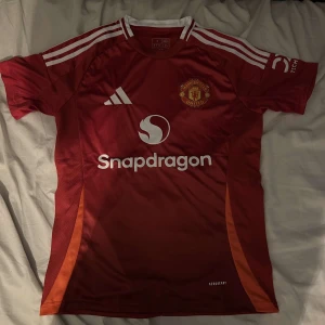 Manchester United hemmatröja 24/25 fotbollströja  - Säljer en röd fotbollströja från Adidas med vita detaljer och klubbmärke på bröstet. Tröjan har korta ärmar och är tillverkad i ett lätt och andningsbart material. Perfekt för fotbollsfans och sportaktiviteter.