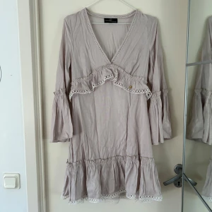 Beige/grå klänning från Yehwang Collection - Säljer en söt beige/grå klänning från Yehwang Collection med volangdetaljer och v-ringning. 