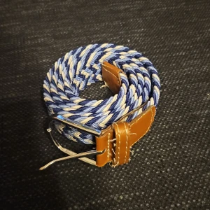 Flätat bälte i blått och beige - Snyggt flätat bälte i blått och beige med bruna skinn-detaljer. Perfekt för att ge en stilren touch till din outfit. Bältet har ett klassiskt spänne i metall och är justerbart för bästa passform. Pris går att diskutera