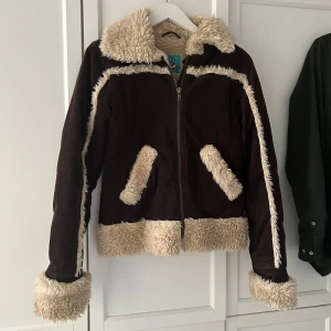 Brun jacka med fuskpälsdetaljer - Cool  brun jacka med mysiga fuskpälsdetaljer i beige. Jackan har dragkedja framtill och är långärmad med fuskpäls vid ärmslut, krage och nederkant. 