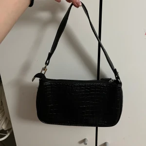 Svart axelväska med krokodilmönster - Snygg svart axelväska med krokodilmönster och dragkedja. Perfekt för att ge en stilren touch till din outfit. Väskan har en justerbar axelrem och är tillverkad i syntetmaterial.