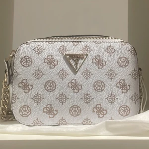 Vit axelväska från Guess - Snygg vit axelväska från Guess med ett elegant mönster i beige. Väskan har två fickor med dragkedja upptill och en justerbar axelrem med kedjedetaljer. Nyskick med prislappen kvar. Dustbag medföljer.
