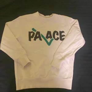 Vit sweatshirt från Palace - Säljer en vit sweatshirt från Palace med ett stort tryck i svart och grönt på framsidan. Tröjan har långa ärmar och en rund halsringning. Perfekt för en avslappnad stil.