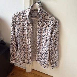 Blommig skjorta med långa ärmar - Snygg blommig skjorta med långa ärmar och klassisk krage. Skjortan har ett färgglatt mönster i nyanser av svart, grått och orange på en vit bakgrund. Perfekt för att liva upp garderoben!