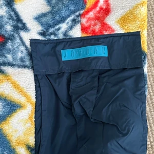 Färgglada fleecebyxor från Jordan - Säljer ett par mjuka och varma fleecebyxor från Jordan med ett färgglatt mönster i rött, gult, blått och vitt. Byxorna har en elastisk midja med snörning och en praktisk ficka på sidan. Perfekta för att hålla sig varm och bekväm.