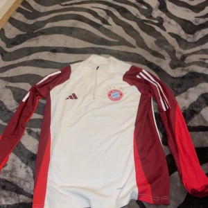 Vit och röd fotbollströja från Adidas - Säljer en snygg vit och röd långärmad fotbollströja från Adidas med FC Bayern München-logga på bröstet. Tröjan har en halv dragkedja framtill och är tillverkad i AEROREADY-material för optimal komfort. Perfekt för fotbollsfans! ⚽