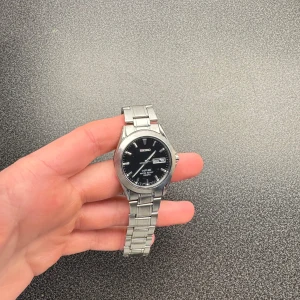 Silverfärgad klocka från Seiko - Snygg och stilren klocka från Seiko med svart urtavla och dag/datumvisning. Boetten och armbandet är i silverfärgat metall. Perfekt för den som gillar en klassisk look. Klockan är kring 20 år gammal och går inte längre att köpa en identisk modell!🤩 Skicket är bra, enbart lite repor på armbandet! Bara att höra av sig vid funderingar🙌🏼