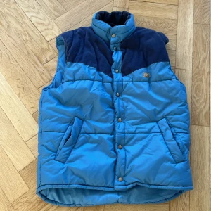 Blå dunväst från Marwin - Säljer en blå dunväst från Marwin med mörkare axelparti och krage. Västen har knappar framtill och två fickor. Perfekt för kyligare dagar. Pris kan diskuteras 