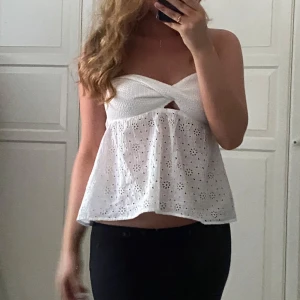 Vit axelbandslös topp - Säljer en söt vit axelbandslös topp med broderade detaljer och en liten knut framtill. Toppen är croppad och har en luftig passform, perfekt för varma dagar. Den har ett fint hålmönster som ger en romantisk känsla. Passar bra till både jeans och kjol.