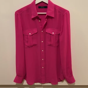 Rosa skjorta från Polo Ralph Lauren - Snygg rosa skjorta från Polo Ralph Lauren med långa ärmar och knappar framtill. Skjortan har två bröstfickor med knappar och är tillverkad i polyester. Perfekt för en färgglad stil!
