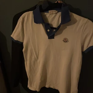 Vit pikétröja från Moncler - Säljer en stilren äkta vit pikétröja från Moncler med blå krage och ärmslut. Tröjan har en klassisk pikédesign med knappar framtill och Monclers logotyp broderad på bröstet. Perfekt för en avslappnad och snygg look. Hör av er vid funderingar. Kan tänka mig och gå ner lite i pris vid en snabb affär