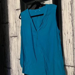 Blå ärmlös blus - Säljer en stilren blå ärmlös blus med v-ringning. Perfekt för varma dagar eller som ett lager under en kavaj. Enkel och elegant design som passar till många tillfällen.