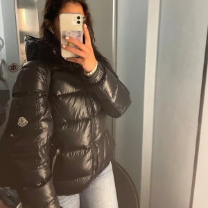 Svart pufferjacka från Moncler - Säljer en snygg svart pufferjacka från Moncler. Jackan har en glansig yta och är perfekt för kyligare dagar. Den har en dragkedja framtill och en hög krage för extra värme 