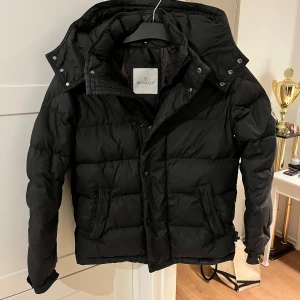 Svart dunjacka från Moncler - Säljer en snygg svart dunjacka från Moncler. Jackan har en pufferdesign med en avtagbar huva och praktiska fickor framtill. Perfekt för kyliga dagar med sin varma och stilrena look.