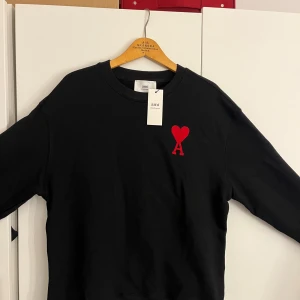 Svart sweatshirt från Ami med rött hjärta - Svart sweatshirt från Ami Alexandre Mattiussi med ikonisk röd broderad hjärtdetalj och bokstaven A på bröstet. Tröjan har rund hals, långa ärmar och en relaxed passform. Tillverkad i mjuk bomull för skön komfort. Perfekt för dig som gillar stilrena plagg med en twist. Priset kan diskuteras privat 