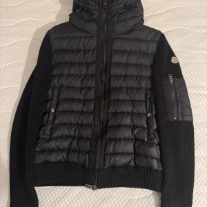 Moncler Cardigan - Svart pufferjacka från Moncler med stickade ärmar och quiltad front. Jackan har huva, dragkedja framtill och Moncler-logga på vänster ärm. 