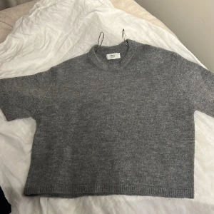 Grå stickad tröja Gina Tricot - Mysig grå stickad tröja från Gina Tricot i ullblandning. Modellen är croppad med korta ärmar och har en rund halsringning. Perfekt för lager-på-lager och passar dig som gillar enkel och stilren look.