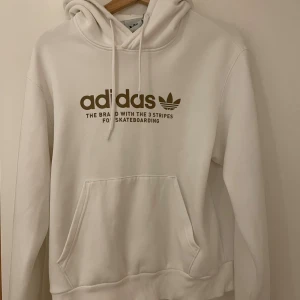 Vit hoodie från Adidas XS - Vit hoodie från Adidas med brunt tryck på bröstet och klassisk logga. Tröjan har huva med snörning, stor magficka och ribbade ärmar. Mjuk och skön i bomullsmix, långärmad och normal passform. Unisex.