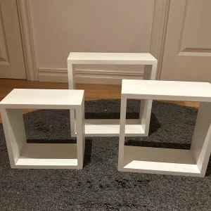 Fina vägghyllor i tre pack säljes - Bilderna visar tre vita hyllor i kubform, perfekta för modern inredning och förvaring. De är i gott skick utan synliga repor eller skador. Passar utmärkt för att visa upp dekorationer, böcker eller småprylar. Stilren design som passar i de flesta hem.