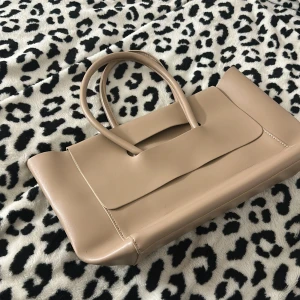 Beige handväska i skinn med stilren design - Snygg beige handväska i skinn med minimalistisk och modern form. Väskan har två handtag och ett stort huvudfack med innerficka. Lock med utskuret handtag ger en cool detalj. Perfekt storlek för allt du behöver och passar till många olika looks.