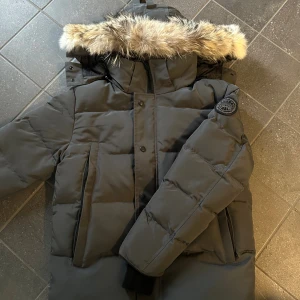 Svart dunjacka Canada Goose med pälskrage - Säljer en svart dunjacka från Canada Goose med klassisk patch på ärmen. Jackan har en stor huva med fluffig pälskant, dragkedja och knappar framtill samt flera praktiska fickor. Perfekt för kalla vinterdagar och riktigt snygg 
