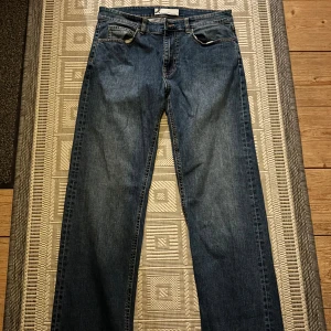 Blå raka jeansbyxor - Klassiska blå jeansbyxor med rak passform och fem fickor. Jeansen har en mörk tvätt och subtila slitningar för en tidlös look. Perfekta för dig som gillar enkel och stilren denim.