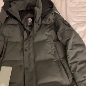 Mörkgrön dunjacka från Canada Goose - Säljer en mörkgrön/grå dunjacka från Canada Goose med klassisk patch på ärmen. Jackan är använd fast i ny skick i storlek S men sitter lite som M