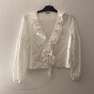 Vit blus med volang från Abby - Säljer en vit blus från Abby i storlek S. Blusen har långa ärmar, v-ringning med volangkant och knyt framtill. Materialet är lätt och har en diskret randig struktur. Perfekt för dig som gillar romantiska detaljer och en feminin look.