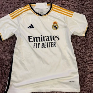 Real Madrid vit matchtröja Adidas S - 🚨MAN KAN BARA TA BORT NAMNET HEMMA🚨Säljer en vit Real Madrid fotbollströja från Adidas i storlek S, slim fit. Tröjan har svarta och gula detaljer, klubbmärke på bröstet, sponsortryck och nummer 7 med namnet Khaleel på ryggen. Tillverkad i lätt och ventilerande polyester med Aeroready-teknologi.