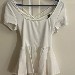 Vit peplumtop med volang och knyt baktill - Supersöt vit peplumtop från Divided med omlott och snörning i ryggen. Toppen har korta ärmar, v-ringad hals och volang nedtill. Aldrig använd.