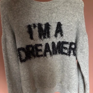 Grå stickad tröja med text - Mysig grå stickad tröja med svart fluffig text 'I'M A DREAMER' framtill. Tröjan har rund halsringning och långa ärmar. Perfekt för dig som gillar statement-plagg och vill ha något unikt och bekvämt i garderoben.