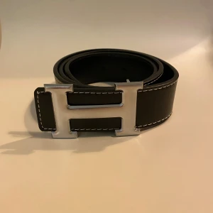 Svart skärp med H-spänne i silver - Stilrent svart bälte med ett stort H-format spänne i silverfärg och vita kontrastsömmar längs kanterna. Tillverkat i skinn och passar perfekt till jeans eller kostymbyxor. Bältet är 100 cm långt och 40 mm brett.