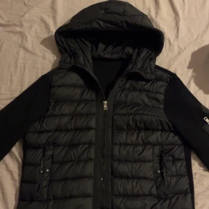 Svart pufferjacka från Moncler - Svart pufferjacka från Moncler med huva och stickade ärmar. Jackan har quiltad framsida, dragkedja framtill och fickor med tryckknappar. På ena ärmen finns en ficka med dragkedja och Moncler-logga. Perfekt för kalla dagar. Pris kan diskuteras!