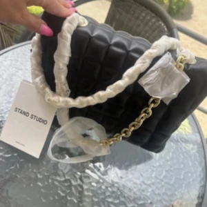 Svart quiltad axelväska Stand Studio - Snygg svart quiltad axelväska från Stand Studio med kedjedetaljer i guld och vadderad design. Väskan har en unik form och ett mjukt material som ger en lyxig känsla. Perfekt för att lyfta din outfit med en cool och modern touch.