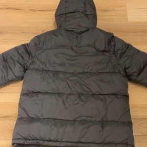 Svart pufferjacka från Polo Ralph Lauren - Svart pufferjacka från Polo Ralph Lauren med huva och flera dragkedjefickor. Jackan har det klassiska röda Polo-logot broderat på bröstet och quiltad design. Perfekt för kalla dagar och har en clean, modern look.