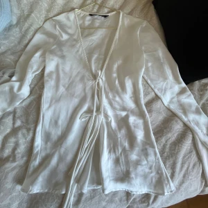 Vit omlottblus med knyt från Zara - Säljer en vit omlottblus från Zara med långa ärmar och v-ringning. Blusen har snygga knytband framtill som ger en cool omlottlook. Materialet är mjukt och har en lätt glansig finish som känns lyxig. 