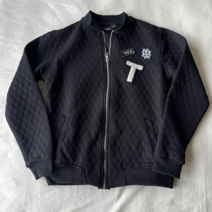 Svart quiltad bomberjacka  - Svart quiltad bomberjacka med dragkedja och applikationer. I storlek 146/152. Fraktar inom 24h.