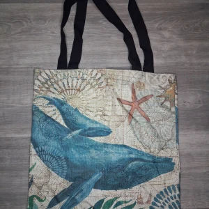 Shoppingväska med blåvalmotiv - Unik shoppingväska med tryck av blåvalar och sjöstjärnor på en beige bakgrund med marina detaljer. Väskan har svarta axelband och är tillverkad i ett tunnare tygmaterial. Perfekt för dig som gillar havsinspirerade accessoarer och vill sticka ut. Mått ca 42x40cm 