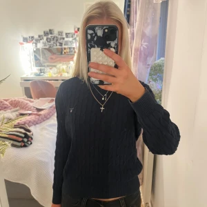 Marinblå kabelstickad tröja GANT - Snygg marinblå kabelstickad tröja från GANT med rund hals och diskret logga på bröstet. Tröjan har långa ärmar och ribbade muddar vid hals, ärmslut och nederkant. Perfekt för dig som gillar klassisk och stilren look.