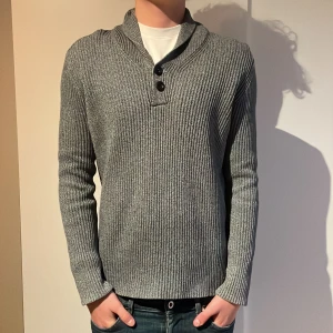 Grå ribbad kofta med knappar - Snygg grå ribbad kofta med krage och två svarta knappar framtill. Tröjan har långärmad passform och är tillverkad i ett stickat material som känns mjukt mot huden. Perfekt för dig som gillar stilrena och enkla plagg med lite extra detaljer vid halsen.