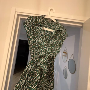 Grön mönstrad långklänning med knyt💚MANGO💚 - Snygg långklänning i grönt med vitt grafiskt mönster. Klänningen har omlottkänsla, knyt i midjan och korta ärmar. Materialet är mjukt och följsamt, perfekt för dig som gillar statement-plagg med coola detaljer.💚