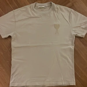 Beige t-shirt från Ami Paris med hjärtdetalj - Snygg beige t-shirt från Ami Paris i bomull. Klassisk passform med korta ärmar och rund hals. På bröstet finns Ami:s ikoniska hjärta i ton-i-ton brodyr. Perfekt för en clean och stilren look.