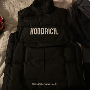 Svart dunväst från Hoodrich - Svart dunväst från Hoodrich med stor vit logga framtill och texten 'FROM NOTHING TO SOMETHING' nedtill. Västen har hög krage, stora fickor och dragkedja i sidan. Perfekt för lager-på-lager och streetwear-stil. Skada vid armhålan