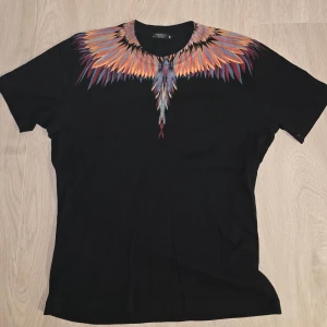 Marcelo Burlon T-shirt - Svart t-shirt från Marcelo Burlon med ikoniskt färgglatt fjädertryck runt halsen i orange, blått och lila. Klassisk passform och korta ärmar. Tillverkad i mjuk bomull, perfekt för dig som vill sticka ut med en unik design.