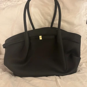 Svart axelväska - Stilren svart axelväska i mjukt skinn med två rejäla handtag och en snygg gulddetalj framtill. Väskan har rymligt brunt innerfoder och stängs med magnetlås. Perfekt för dig som vill ha en klassisk och tidlös look. Den är aldrig använd💕