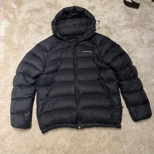 Svart dunjacka från Peak Performance - Säljer en svartdunjacka från Peak Performance i strl L, modell  M Down Hood Jacket FW23. Jackan har huva med dragsko och dragkedja framtill. Perfekt för kalla dagar med sin puffiga look och stilrena svarta färg. Passar dig som gillar funktion och enkel stil.