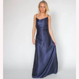 Marinblå långklänning By Stina - Säljer en elegant marinblå långklänning med v-ringad urringning och smala axelband. Klänningen har en stilren, figurnära passform och är tillverkad i ett glansigt material som ger en lyxig känsla. Perfekt för bal eller student. Snygg och tidlös design!