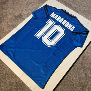 Argentina Maradona 10 Adidas tröja blå - Retro Argentina fotbollströja från Adidas i blått med svarta detaljer, klassiskt nummer 10 och Maradona på ryggen. Tröjan har korta ärmar, krage med knapp och broderat landslagsemblem. Materialet är glansigt och lätt, perfekt för fotbollsfans.