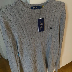 Grå stickad tröja från Polo Ralph Lauren - Säljer en stilren grå stickad tröja från Polo Ralph Lauren. Tröjan har ett klassiskt flätmönster och den ikoniska loggan broderad på bröstet. Perfekt för en avslappnad men ändå elegant look.  Taggen följer inte med.