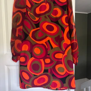 Retro kortklänning eller tunika med färgstarkt mönster - Färgstark kortklänning eller tunika med retroinspirerat, abstrakt mönster i orange, rött och olivgrönt. Klänningen har långa, transparenta ärmar och en rak passform. Perfekt för dig som vill sticka ut med en unik look. Materialet känns lätt och luftigt.